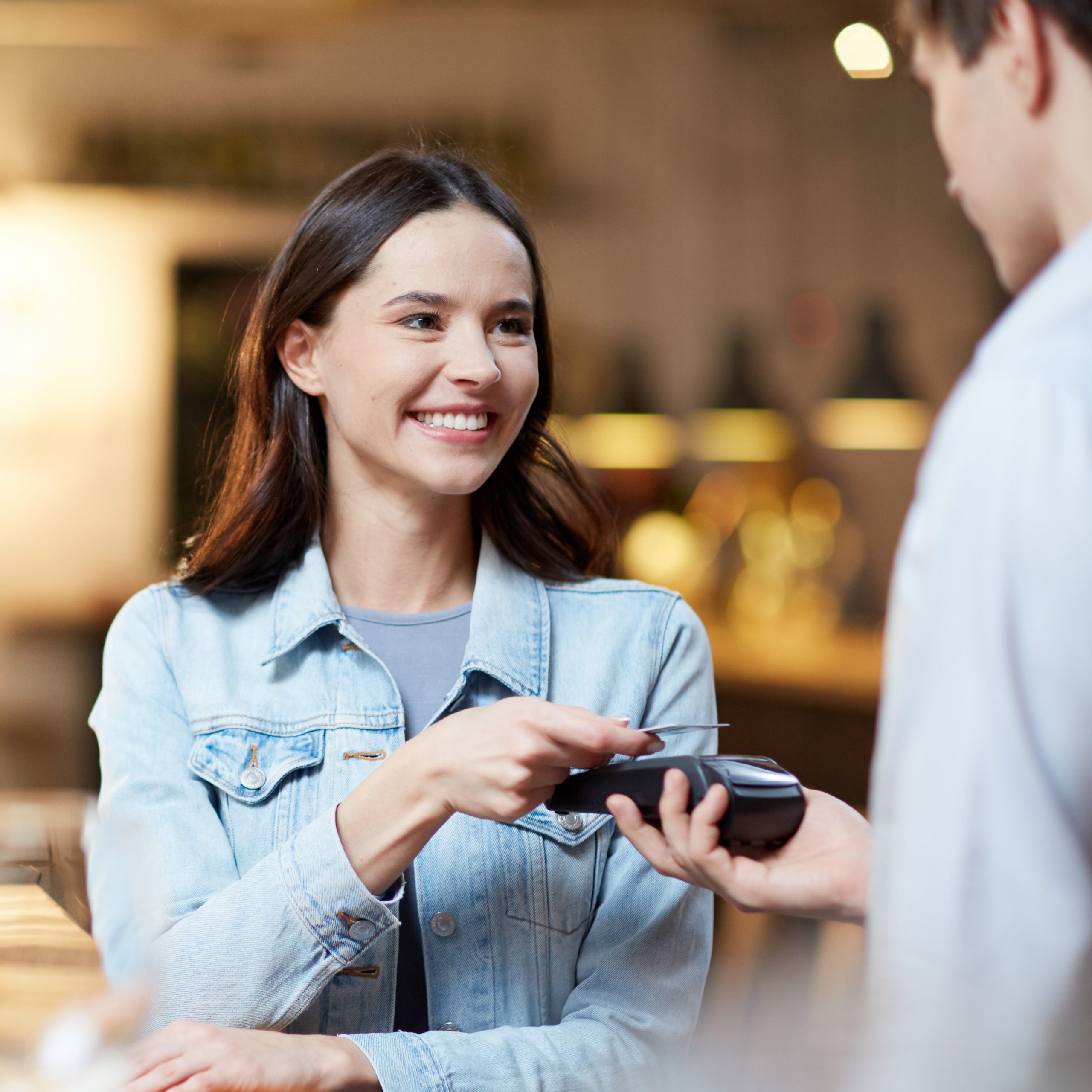 Young smiling woman with card holding it over payment terminal while paying for her order in cafe